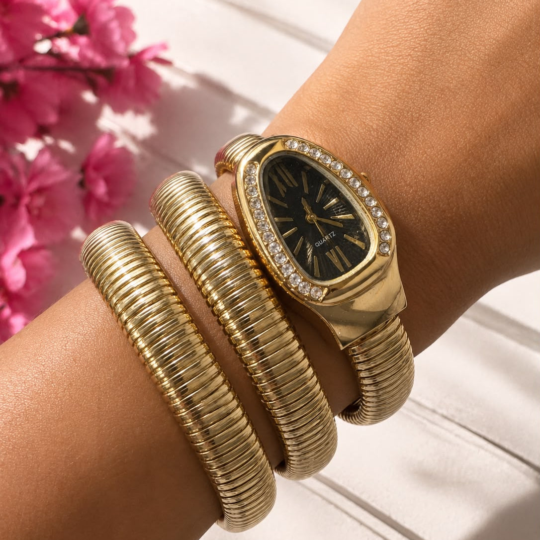 A gold-tone watch with a black dial and crystal-encrusted bezel, styled on a wrist alongside a matching gold bangle bracelet, with pink flowers in the background.