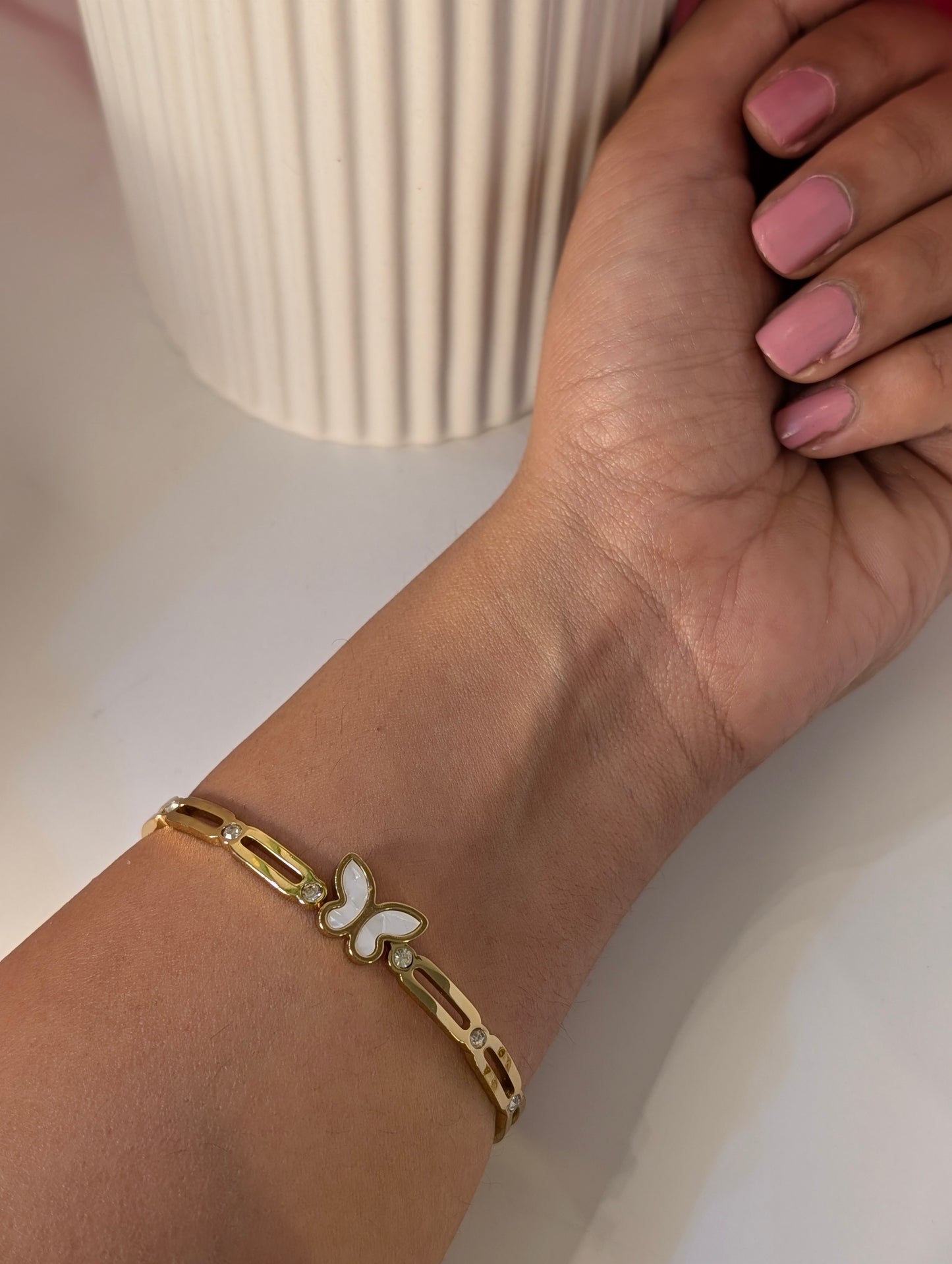 Gold bracelet with a butterfly charm on a wrist, against a neutral background