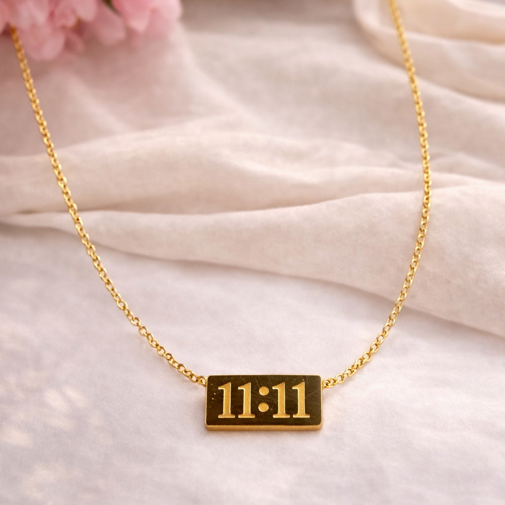 Gold necklace with a rectangular pendant displaying '11:11' on a soft pink fabric background.