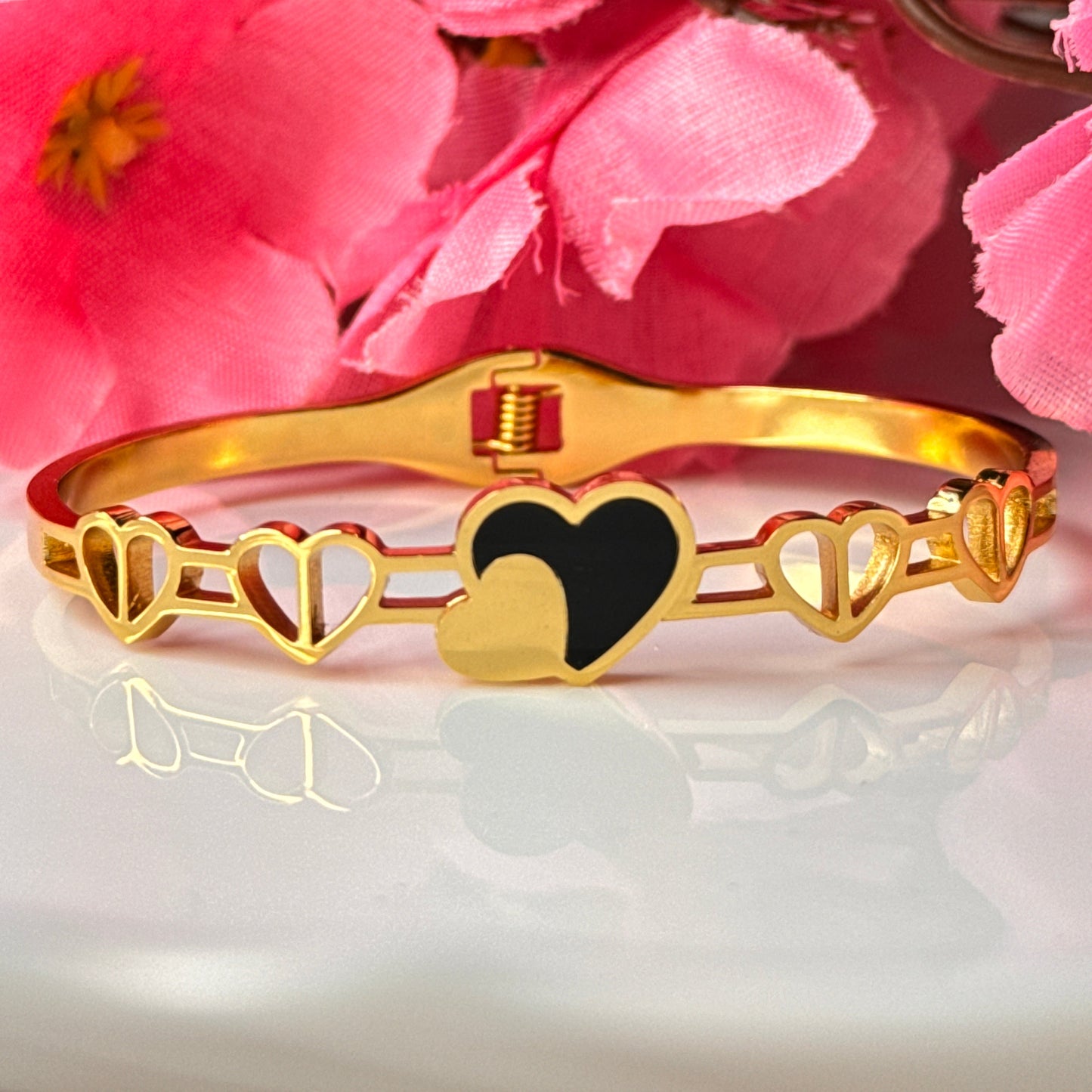 A gold-colored bracelet with a black heart charm, displayed against a backdrop with pink flowers.