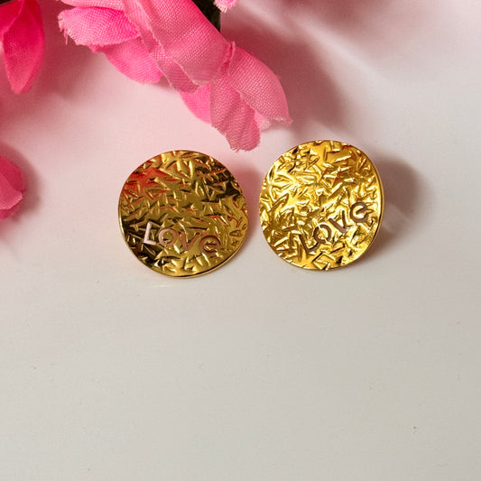 A pair of gold stud earrings with a textured surface, displayed against a white background with pink floral accents.