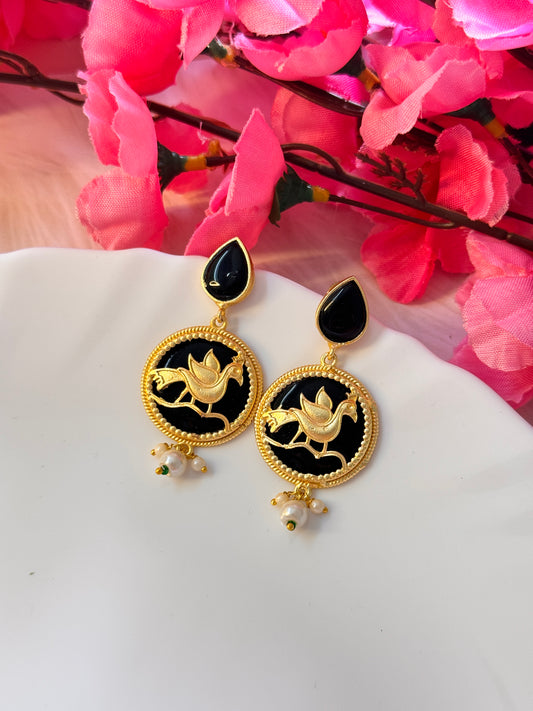 A pair of gold-colored peacock earrings with black and vibrant green details, displayed against a floral background.