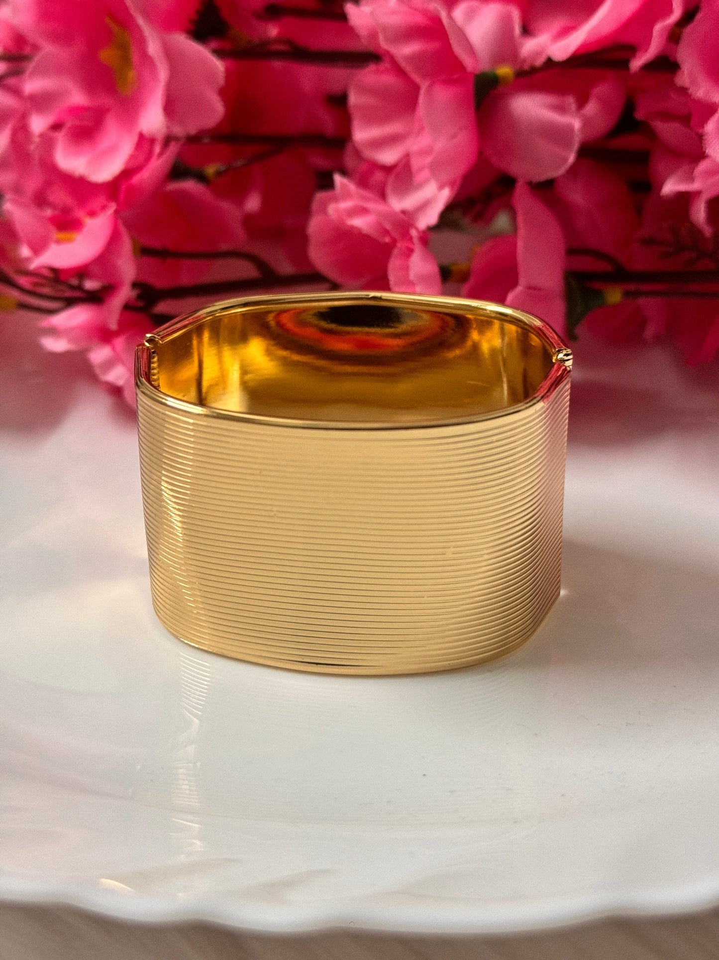 A gold plated cuff bracelet displayed on a white surface with pink floral background.