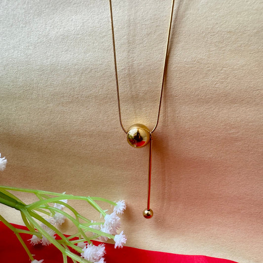 A gold-colored necklace with a simple design placed on a beige background with some floral designs to the side.