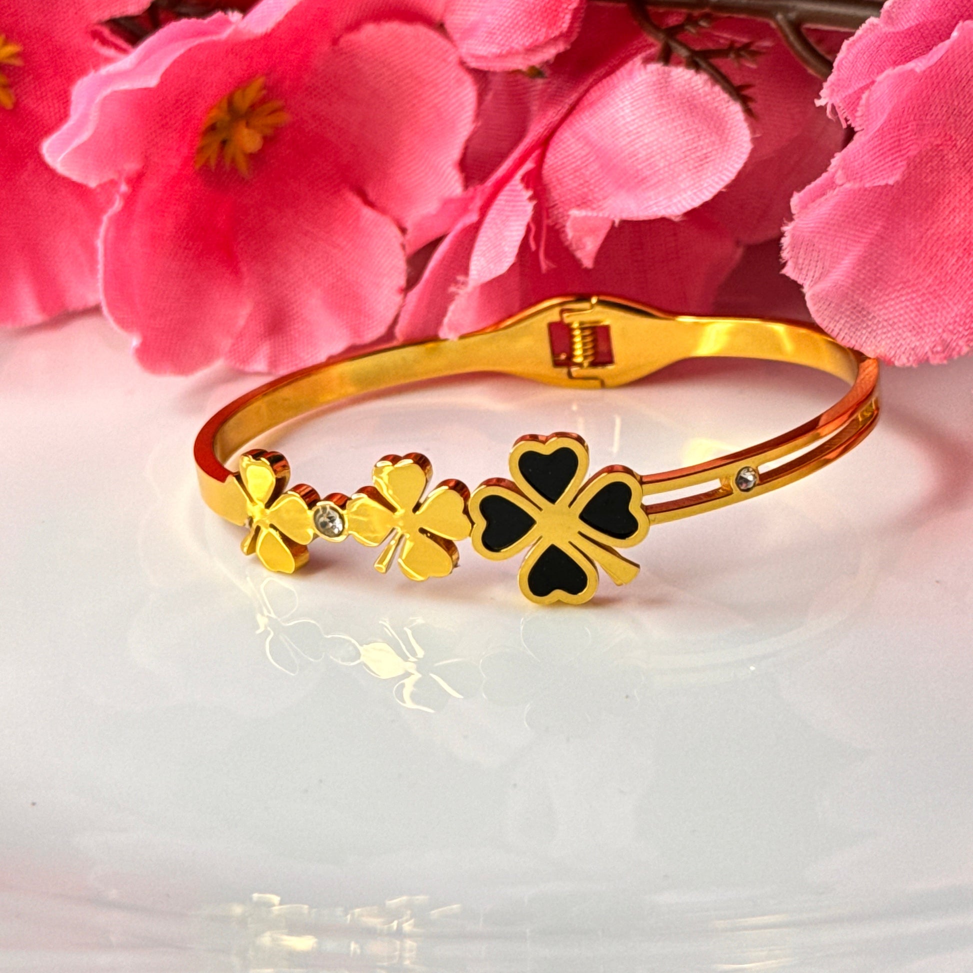 A gold-plated bracelet with clover designs displayed on a white surface with pink flowers in the background.