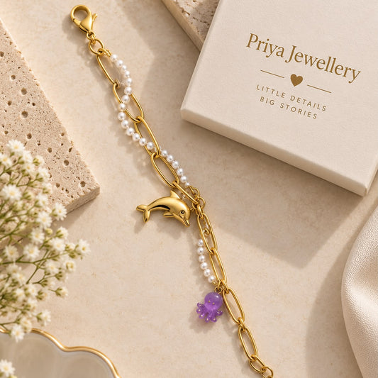 A gold chain bracelet with white pearl-like beads, a dolphin charm, and a purple elephant charm, styled on a beige surface with a Priya Jewellery box and dried flowers.