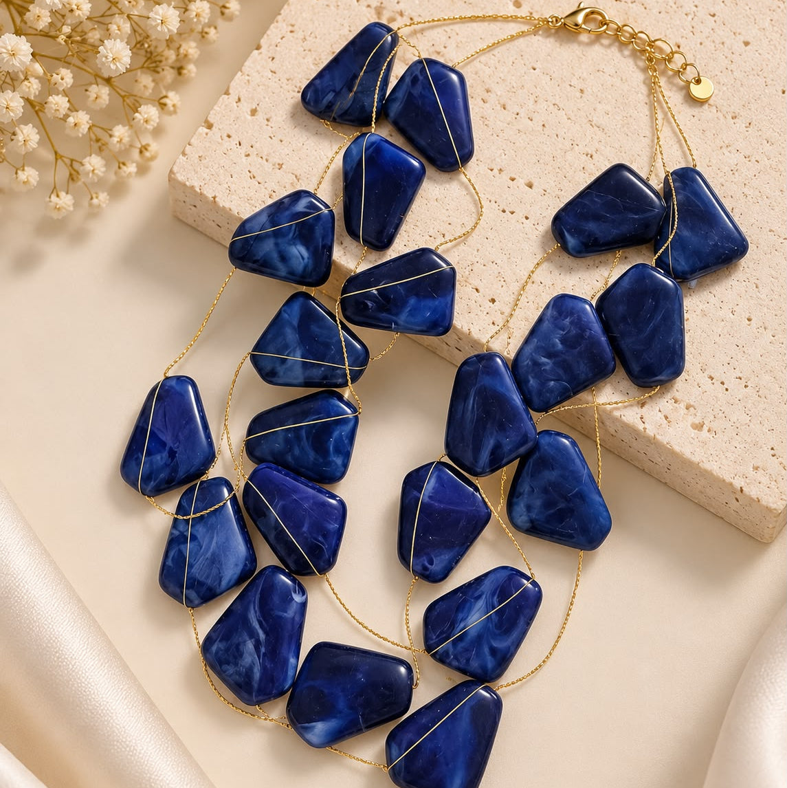 A multi-strand statement necklace featuring deep blue marbled stone chips on a gold chain, styled on a beige textured stone slab with cream satin and dried flowers.