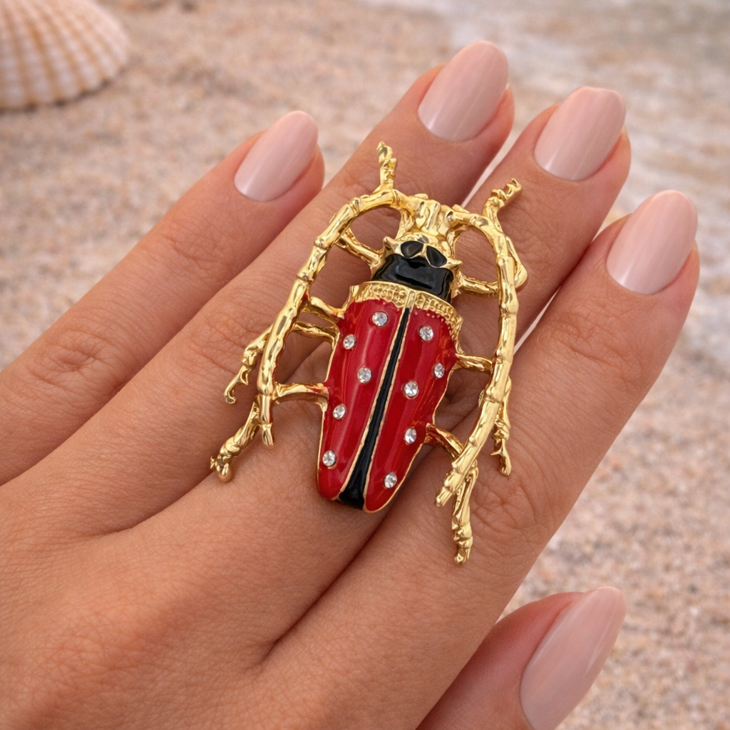Red and gold beetle-shaped ring on a hand with a beach background