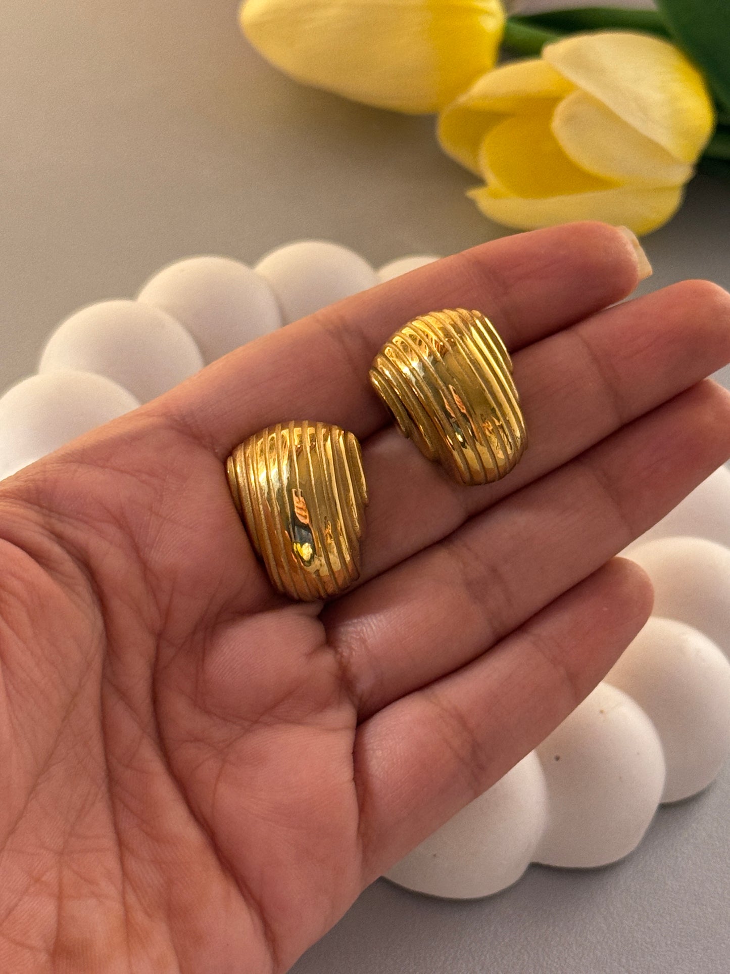 Gold earrings held in a hand with yellow tulips and white stones in the background
