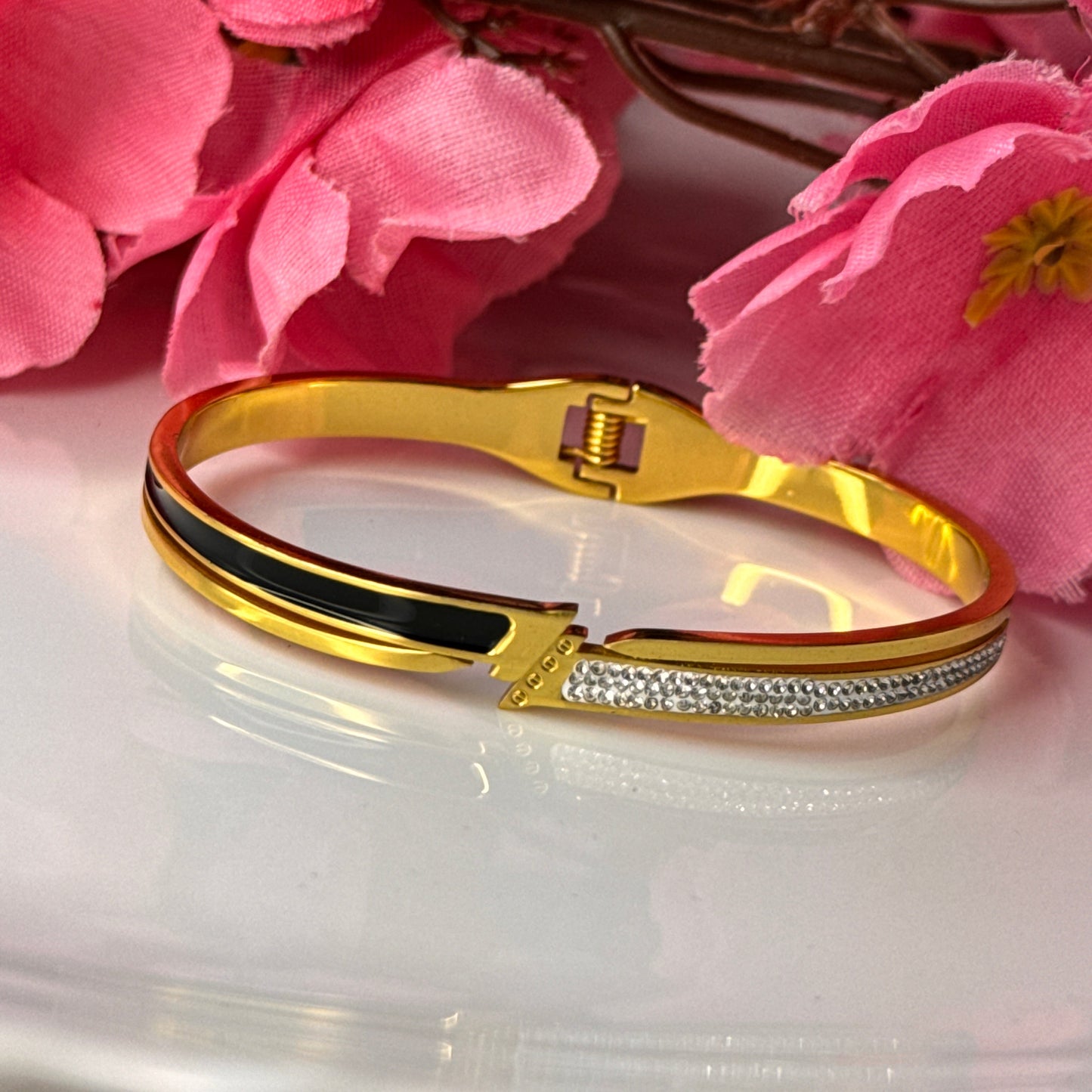 A gold bracelet with a black and white pattern, featuring a clasp, displayed on a white surface with pink flowers in the background.