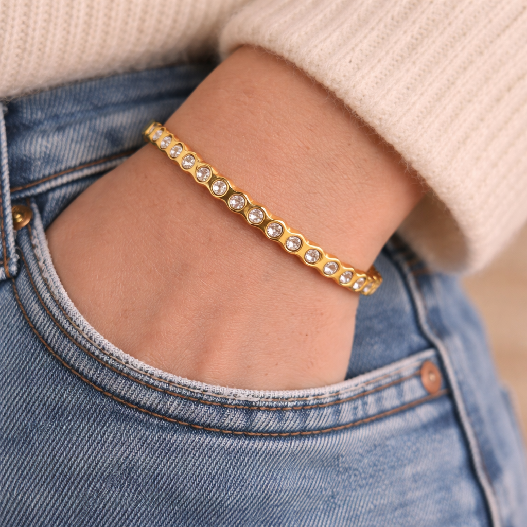 Gold bracelet with clear stones on a wrist, wearing a beige sweater and blue jeans.