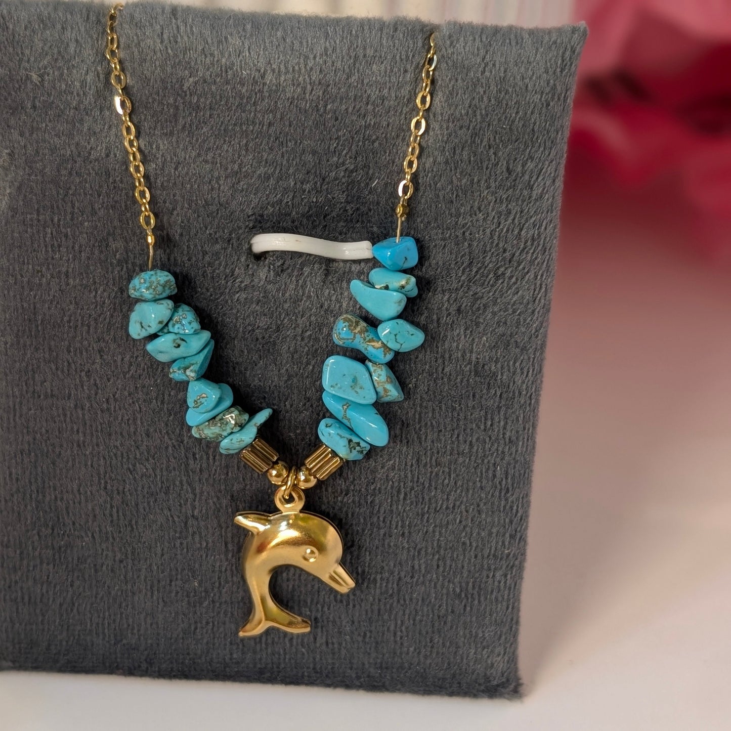 Necklace with turquoise beads and a gold dolphin charm on a gray stand.