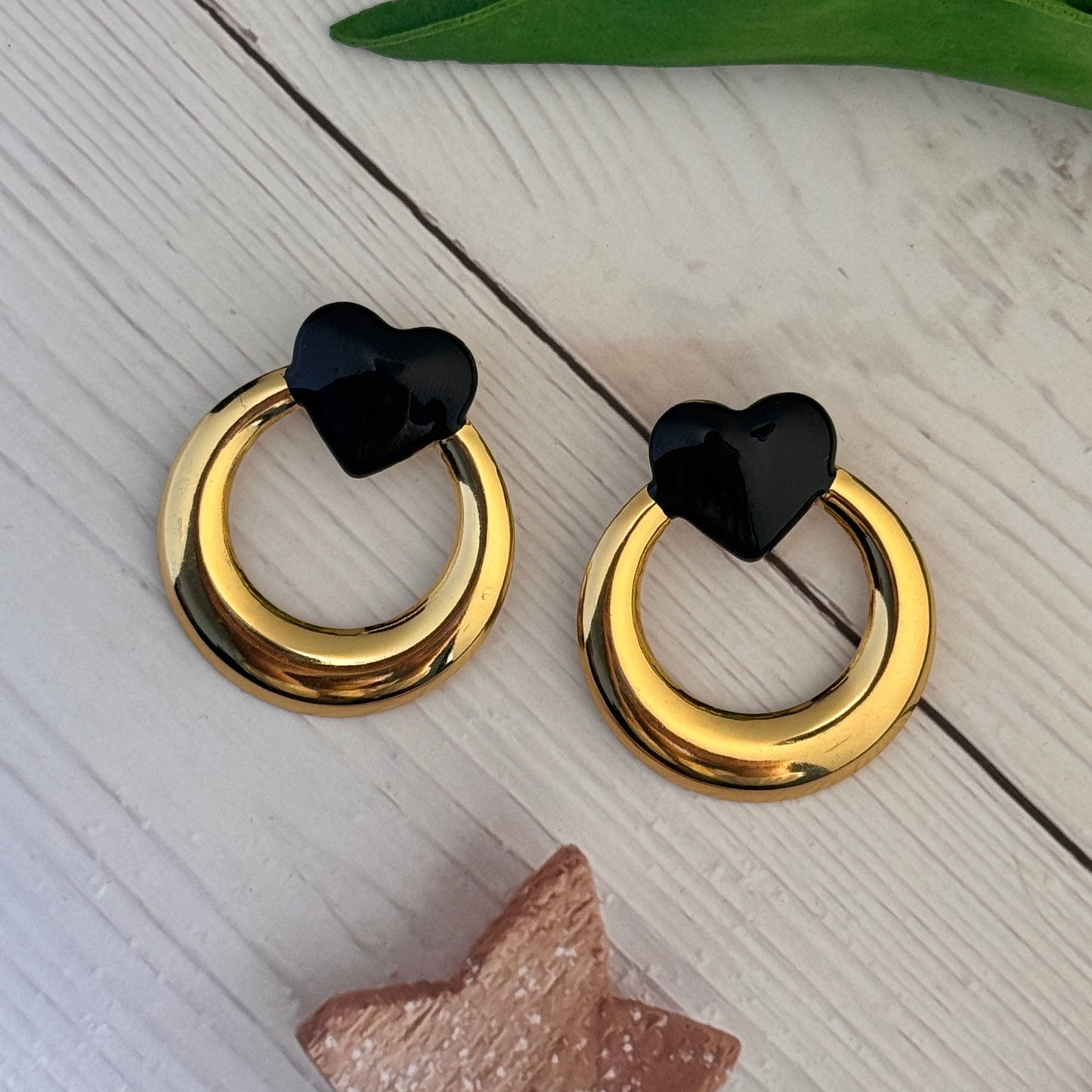 Gold hoop earrings with black heart-shaped ends on a light wooden surface.