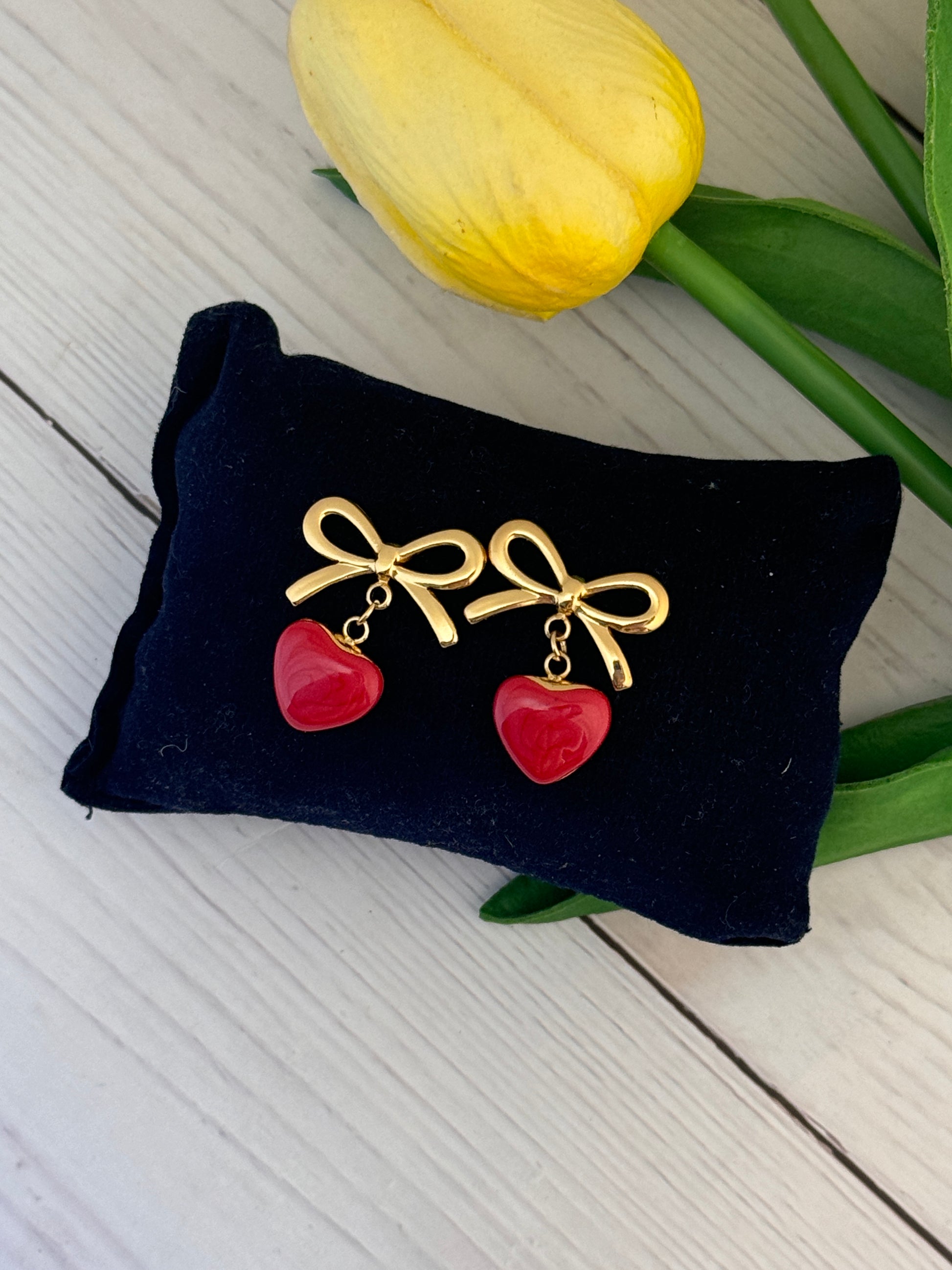 Gold bow and red heart earrings on a black pillow with a yellow tulip in the background.