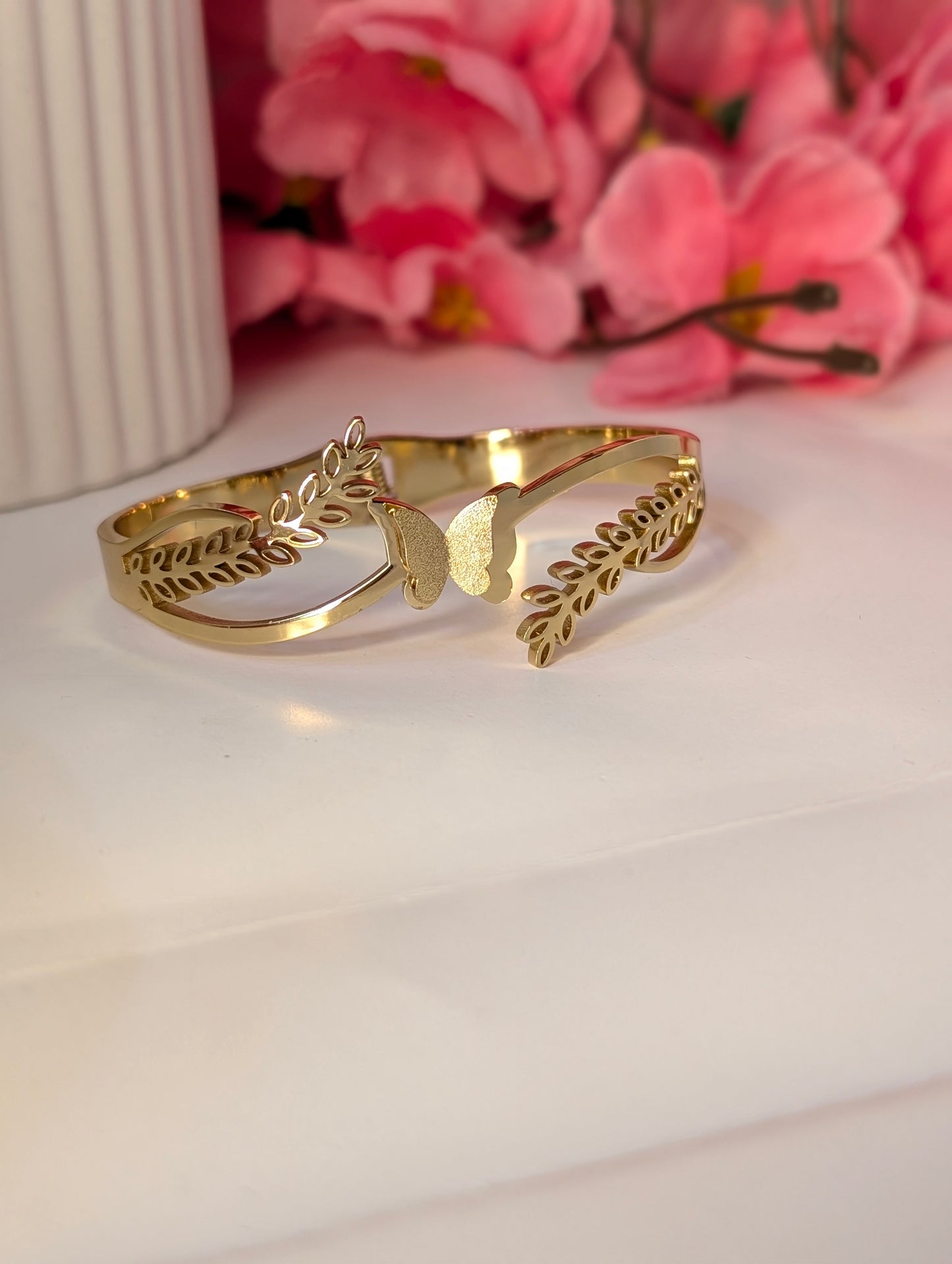 Gold bracelet with leaf design on a white surface with pink flowers in the background