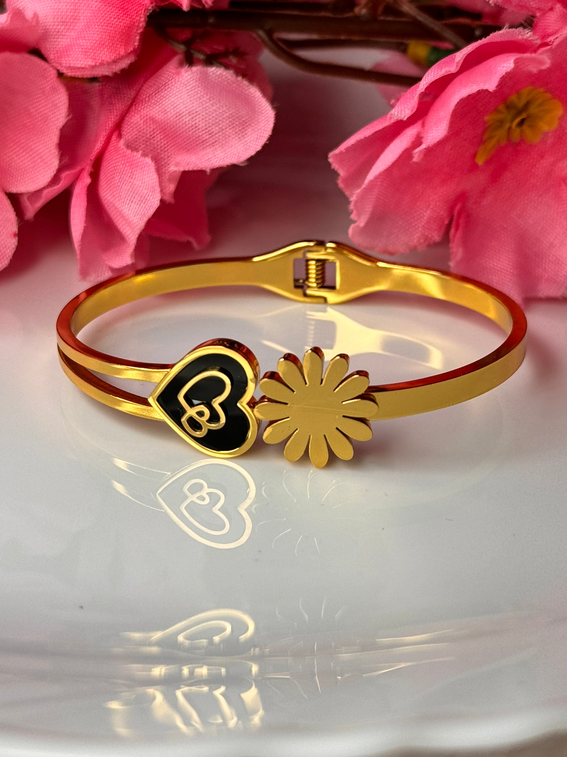 A gold-colored bracelet with a floral design and a heart charm, displayed on a white surface with pink flowers in the background.