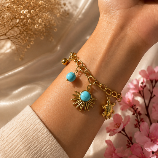 A gold chain charm bracelet with turquoise and sunburst charms, shown worn on a woman's wrist. The background features a draped beige fabric, dried flowers, and pink blossoms.