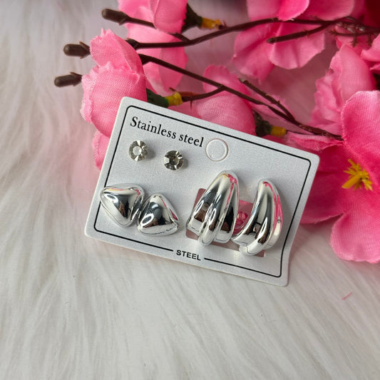 A set of three silver stud earrings on a white card with 'Stainless steel' text, displayed alongside pink flowers.