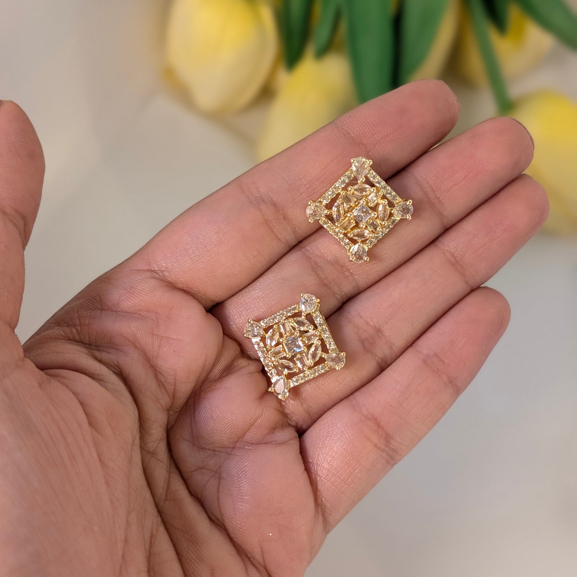 Gold square-shaped earrings held in a hand with a blurred floral background