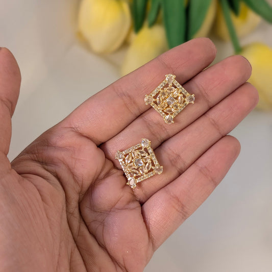 Gold square-shaped earrings held in a hand with a blurred floral background