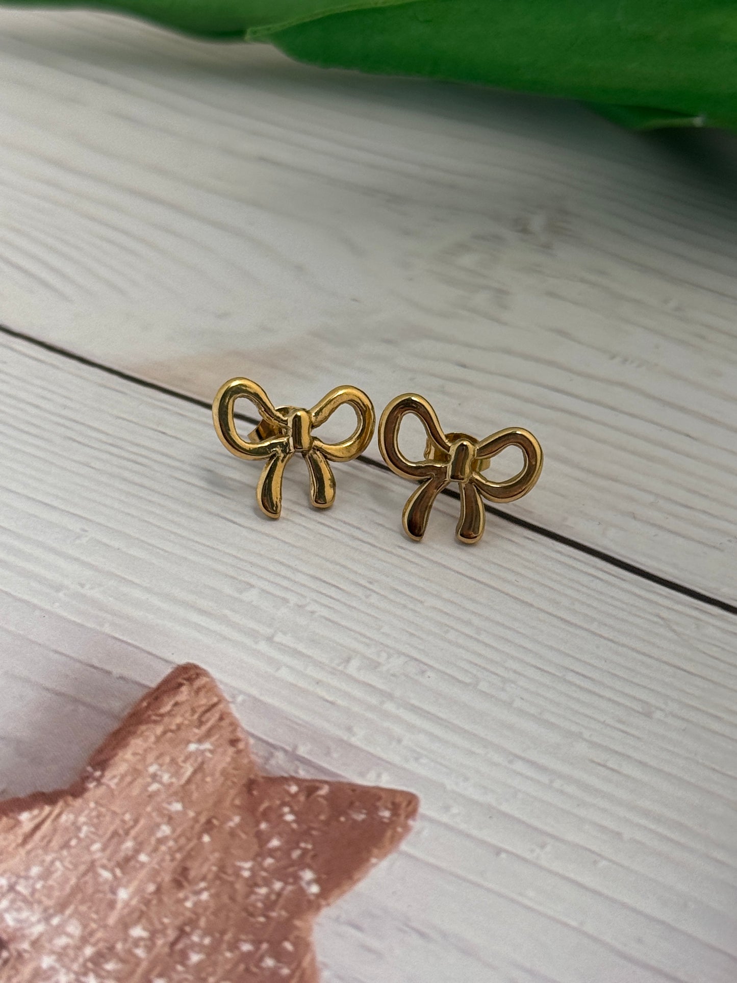 Gold bow-shaped earrings on a light wooden surface with a pink star and green leaf in the background.