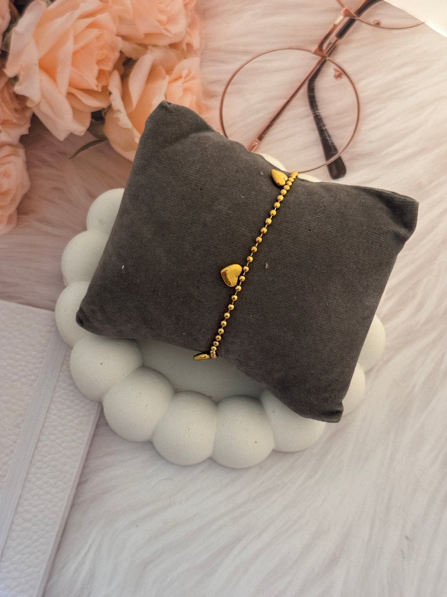 Gold bracelet with heart charms on a gray pillow, surrounded by flowers and glasses.
