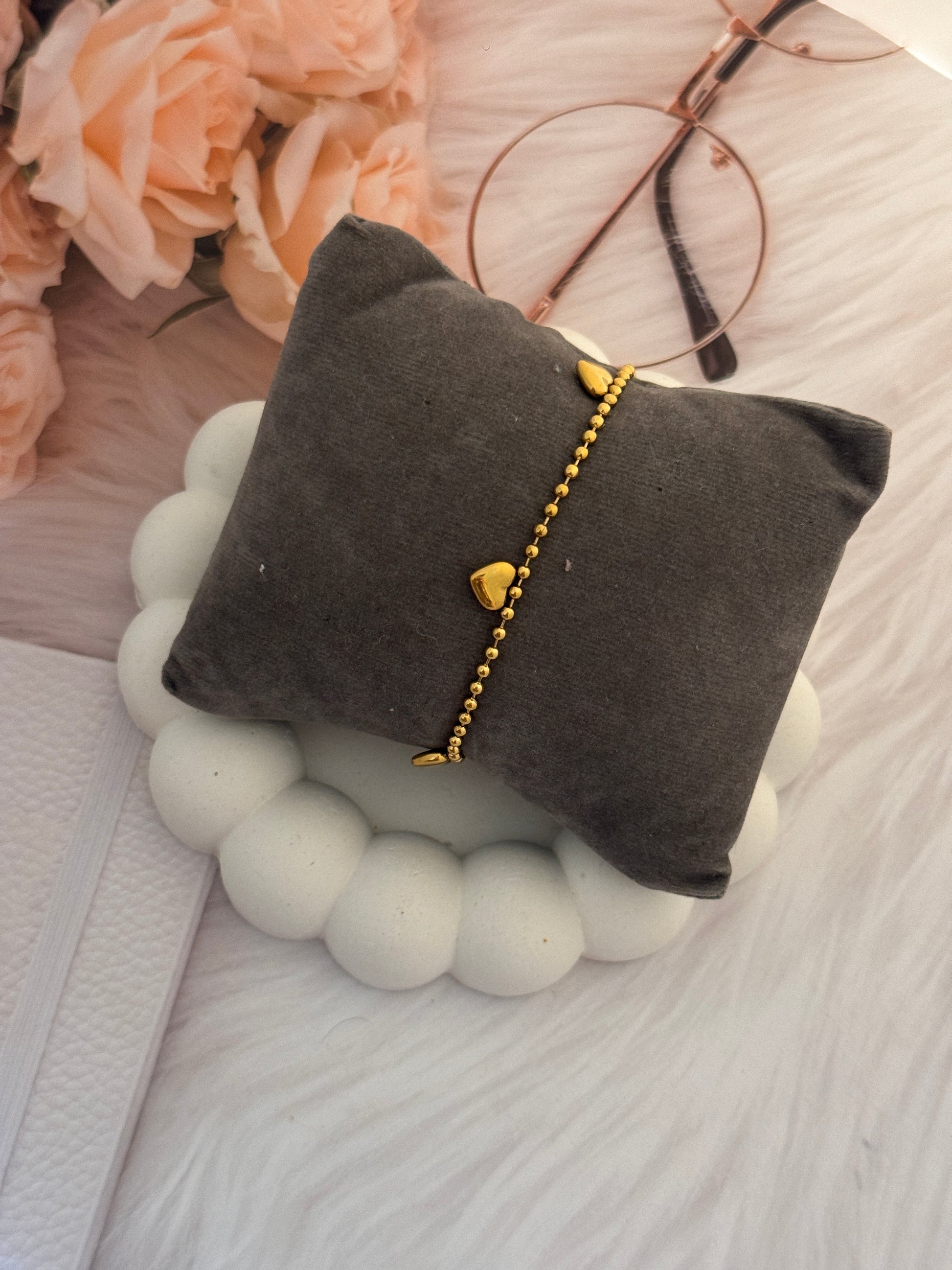 Gold bracelet with heart charms on a gray pillow, surrounded by flowers and glasses.