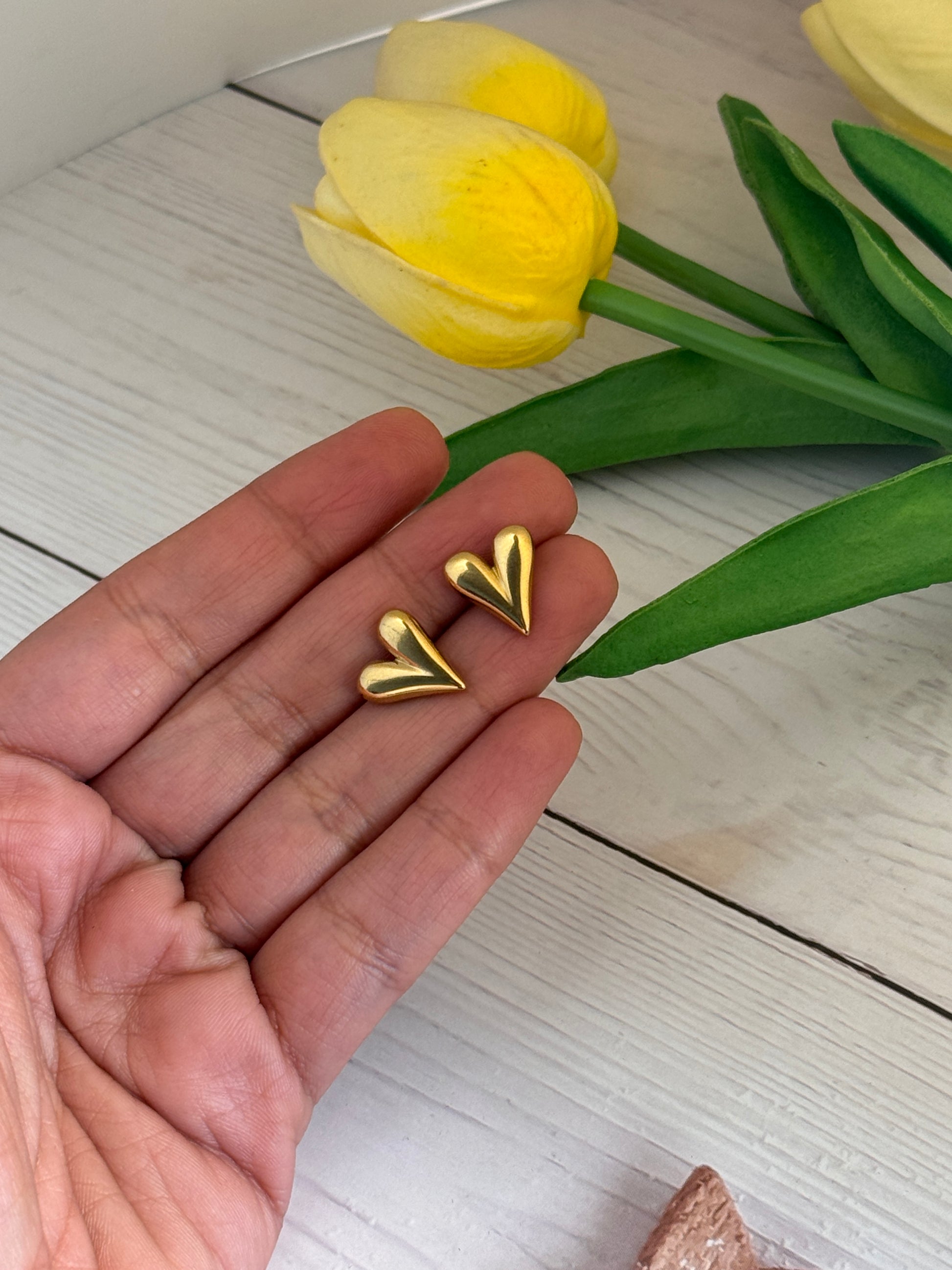 Gold heart-shaped earrings held in a hand with yellow tulips on a light wooden surface