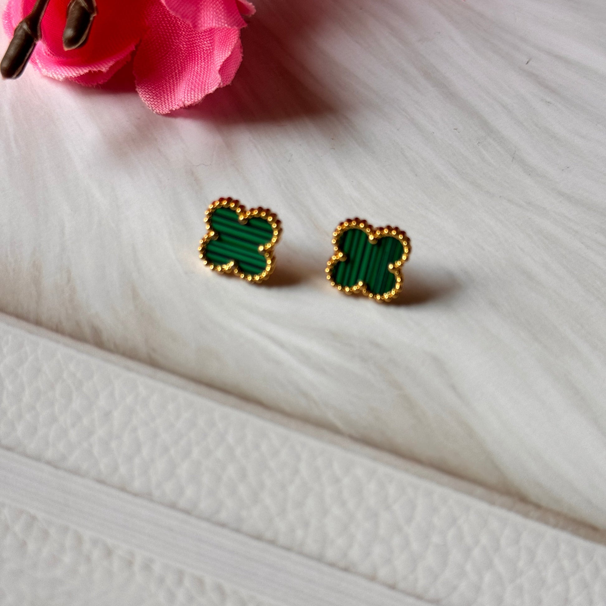 A pair of gold and green clover earrings displayed on a white surface with a pink flower in the background.