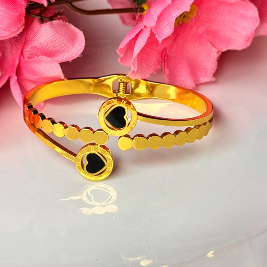 A gold-colored bracelet with a heart shape and a black stone in the center, displayed against a floral background.