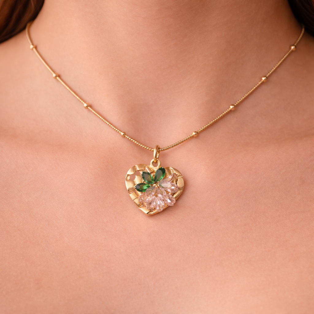 Gold necklace with a heart-shaped pendant featuring green and pink stones on a person's neck.