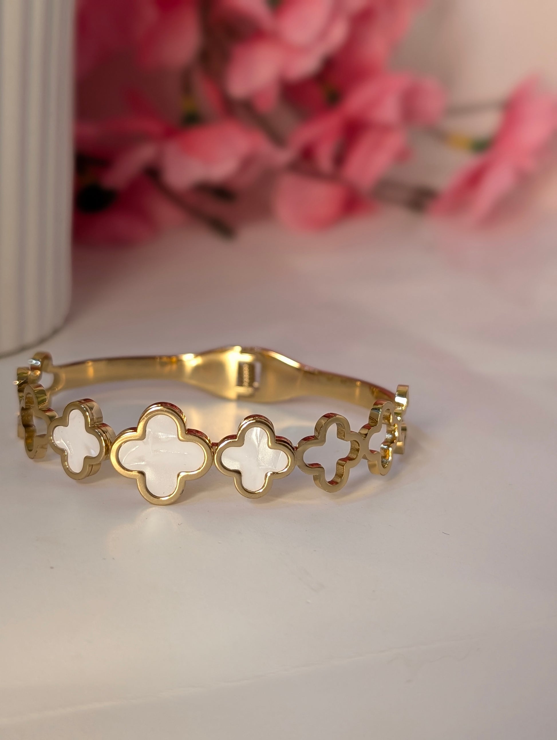 Gold bracelet with white clover-shaped charms on a light surface with pink flowers in the background