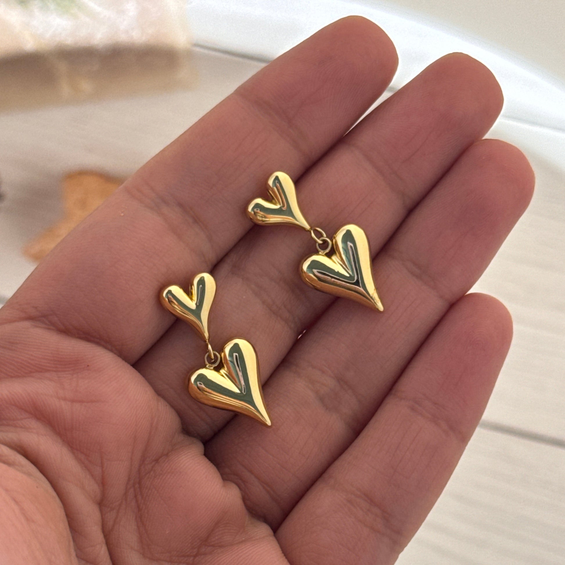 Gold heart-shaped earrings held in a hand against a neutral background