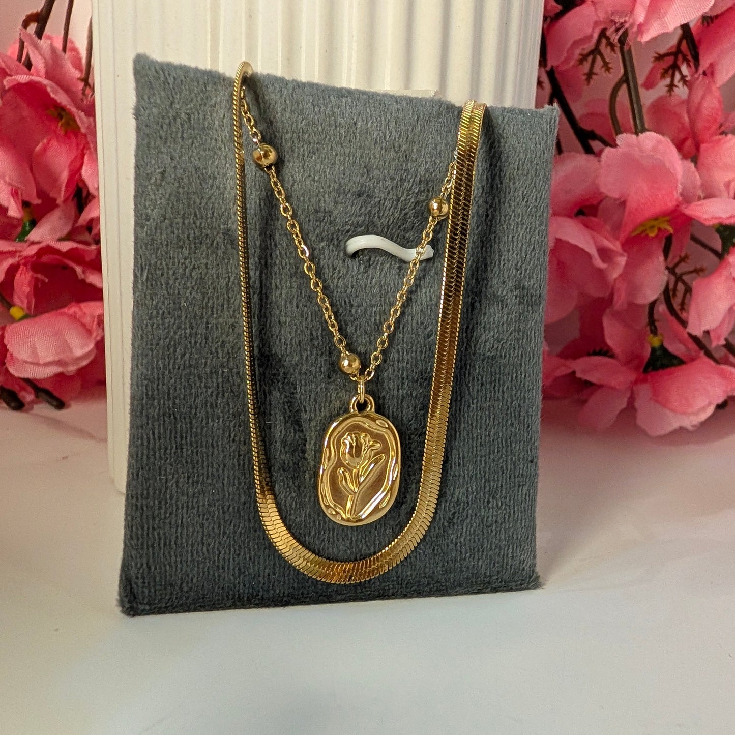 Gold necklace with a pendant on a gray jewelry pillow with pink flowers in the background