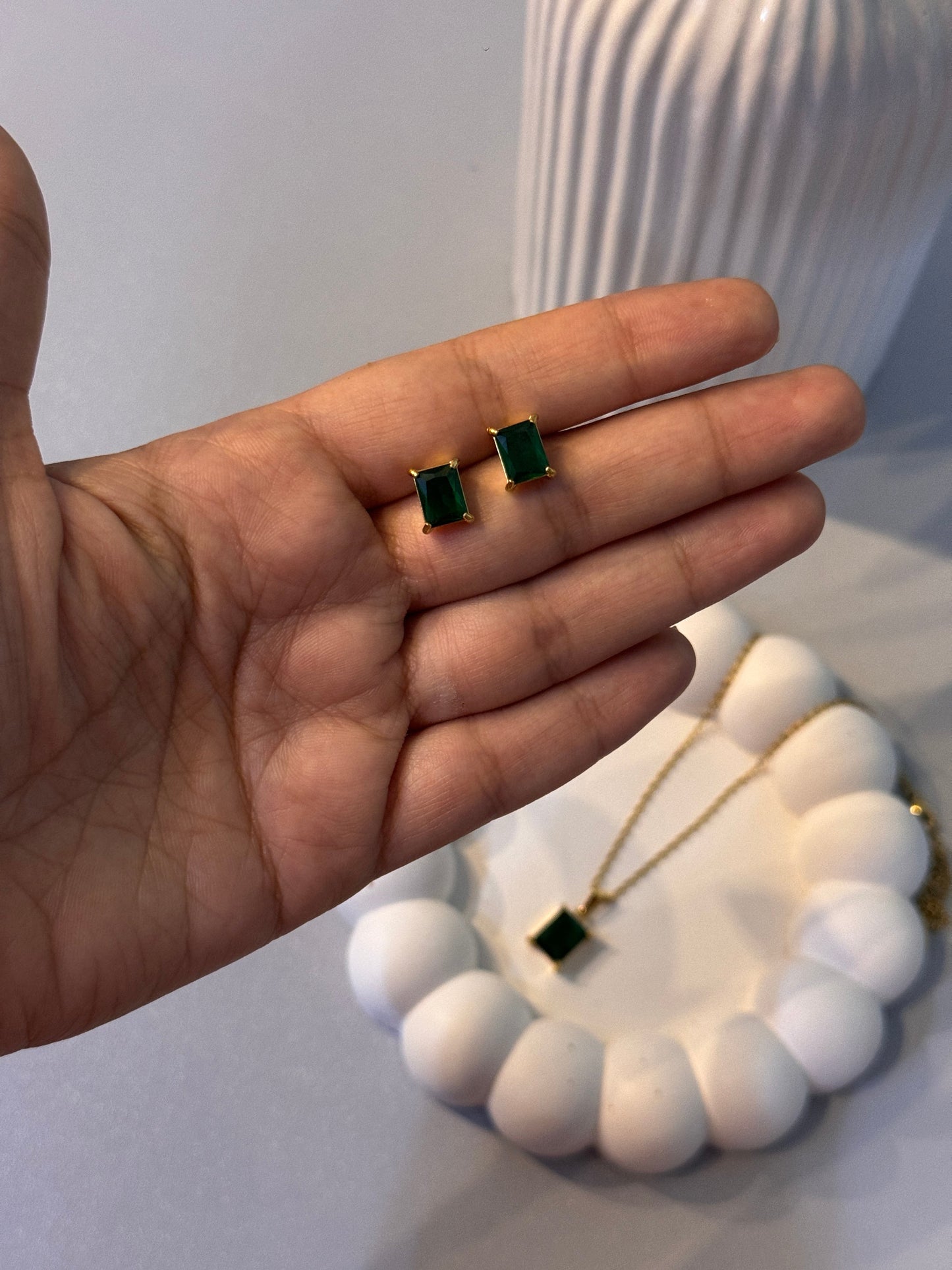 Two green square earrings held between fingers with a blurred white decorative object in the background.