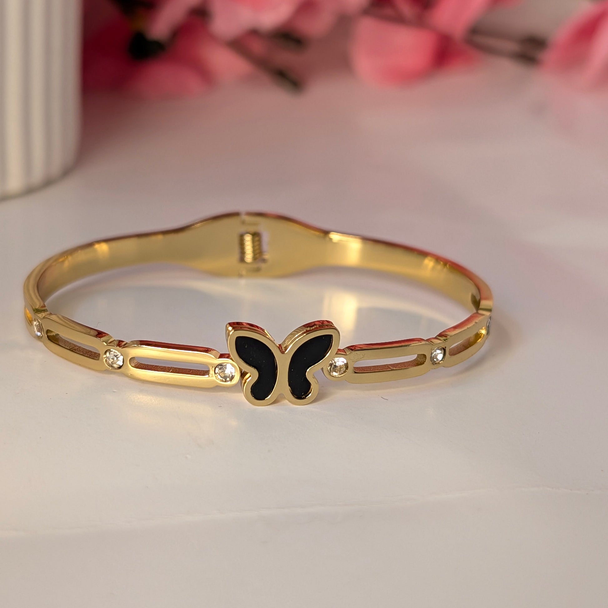 Gold bracelet with a butterfly charm on a light surface with pink flowers in the background
