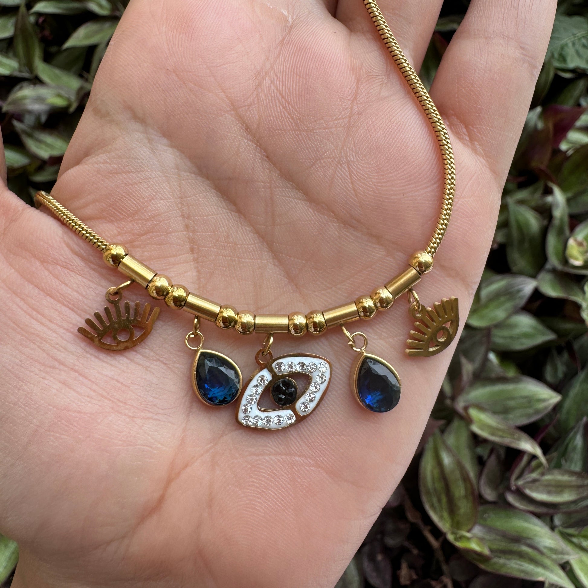 Gold bracelet with heart and blue gemstone charms held in a hand against a green leafy background