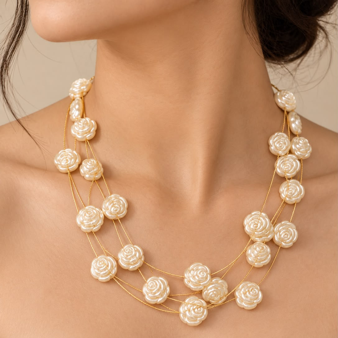 A close-up of a model wearing a multi-strand necklace with cream-colored rose-shaped beads on a gold chain, paired with a white off-the-shoulder top.
