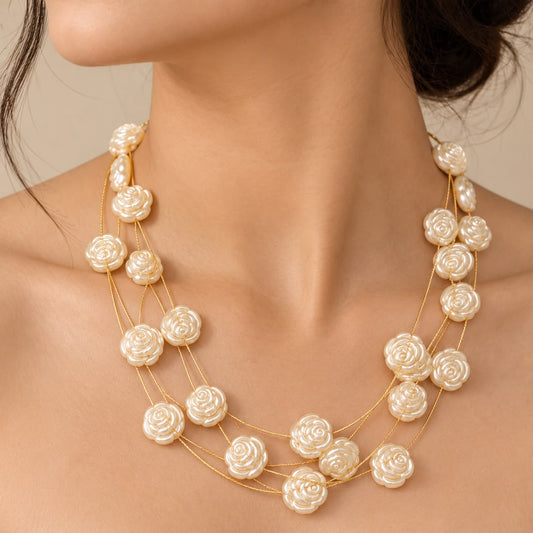 A close-up of a model wearing a multi-strand necklace with cream-colored rose-shaped beads on a gold chain, paired with a white off-the-shoulder top.