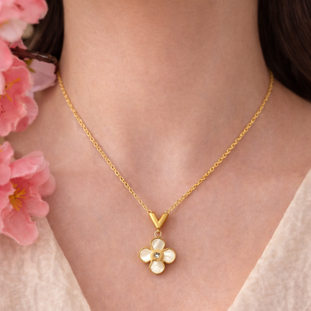 Gold necklace with a floral pendant worn by a person, with pink flowers in the background.