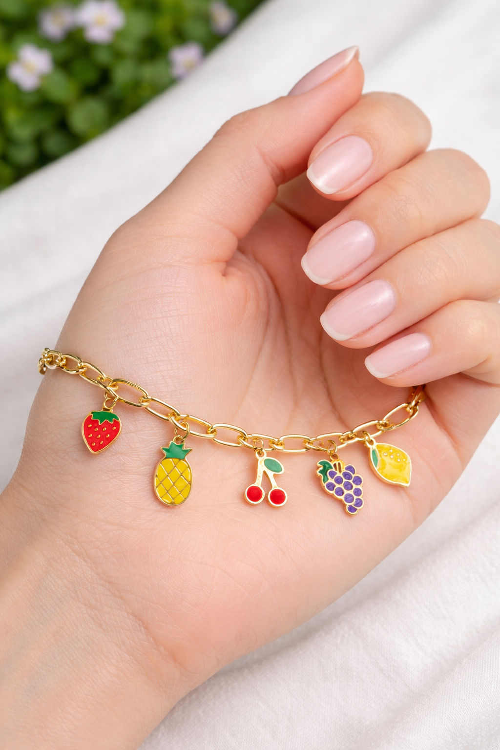 Gold bracelet with colorful fruit charms held by a hand with pink nail polish.