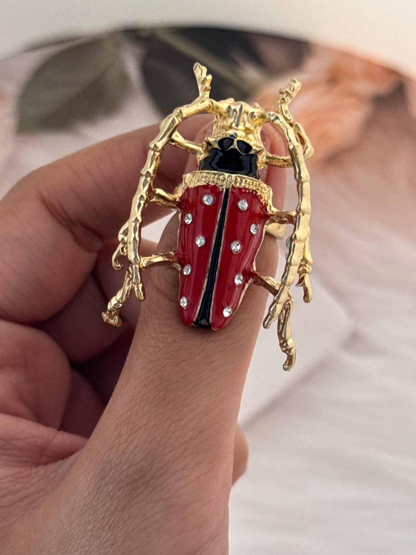Red and gold beetle-shaped brooch held by a hand against a neutral background