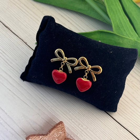 Gold bow earrings with red heart charms on a black pillow with green leaves and a star decoration in the background.