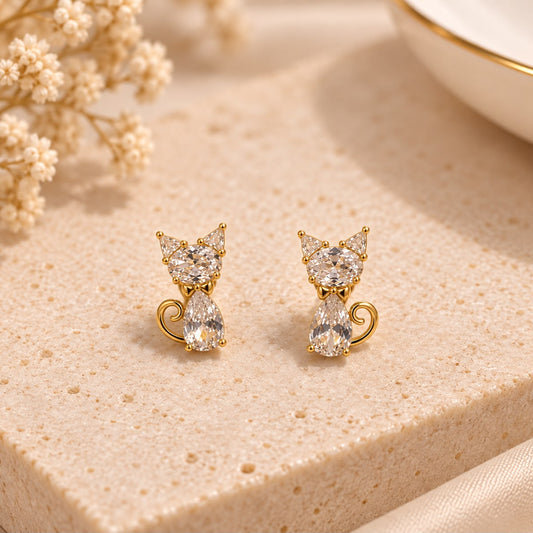 A pair of gold cat-shaped stud earrings with clear teardrop gemstones, displayed on a textured beige stone surface with dried flowers in the background.