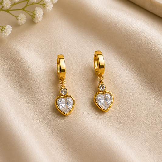 Gold heart-shaped earrings with clear stones on a beige fabric background