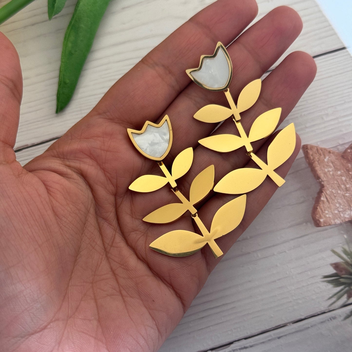 Gold leaf-shaped earrings held in a hand on a light wooden surface.