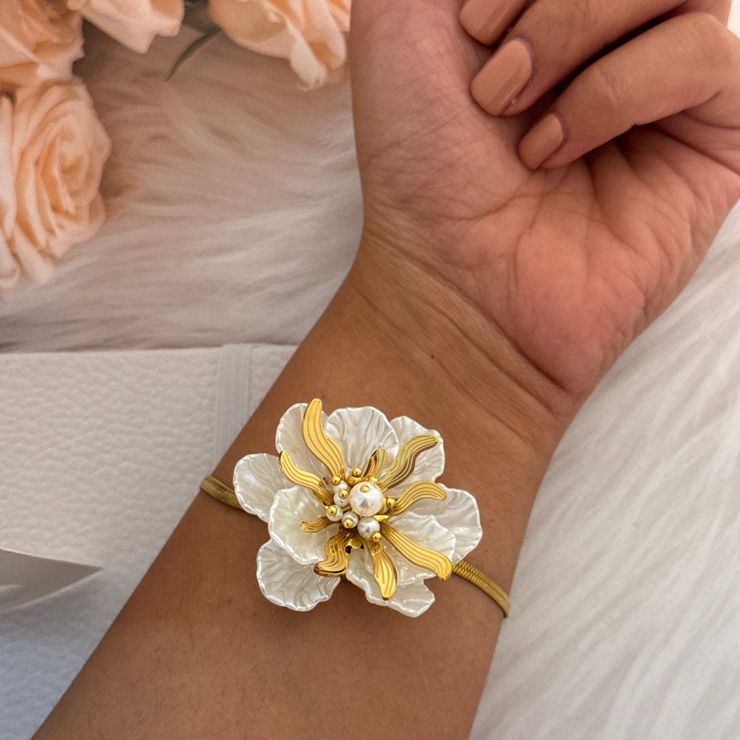 Gold and white floral bracelet on a wrist with a blurred background of flowers and fabric.