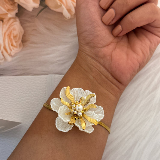 Gold and white floral bracelet on a wrist with a blurred background of flowers and fabric.