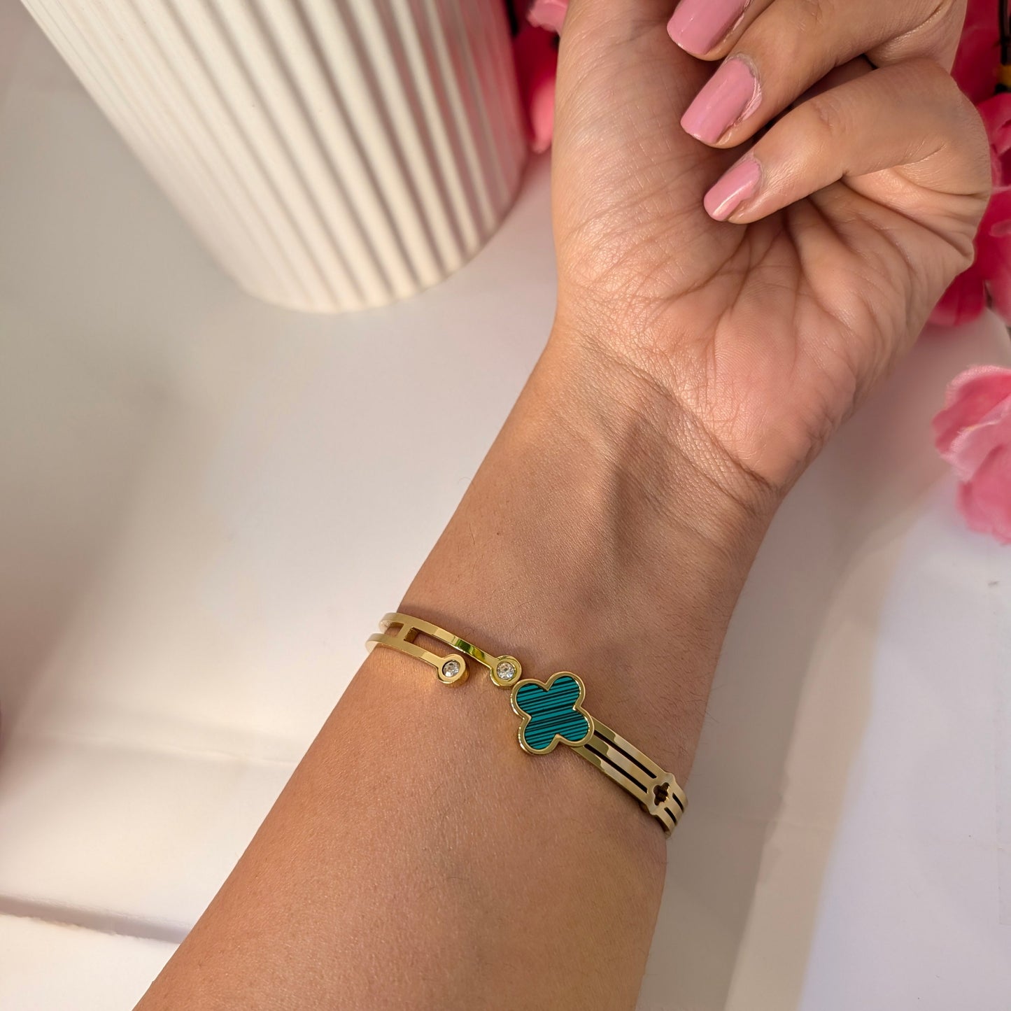 Gold bracelet with a green heart charm on a wrist, against a soft pink floral background.