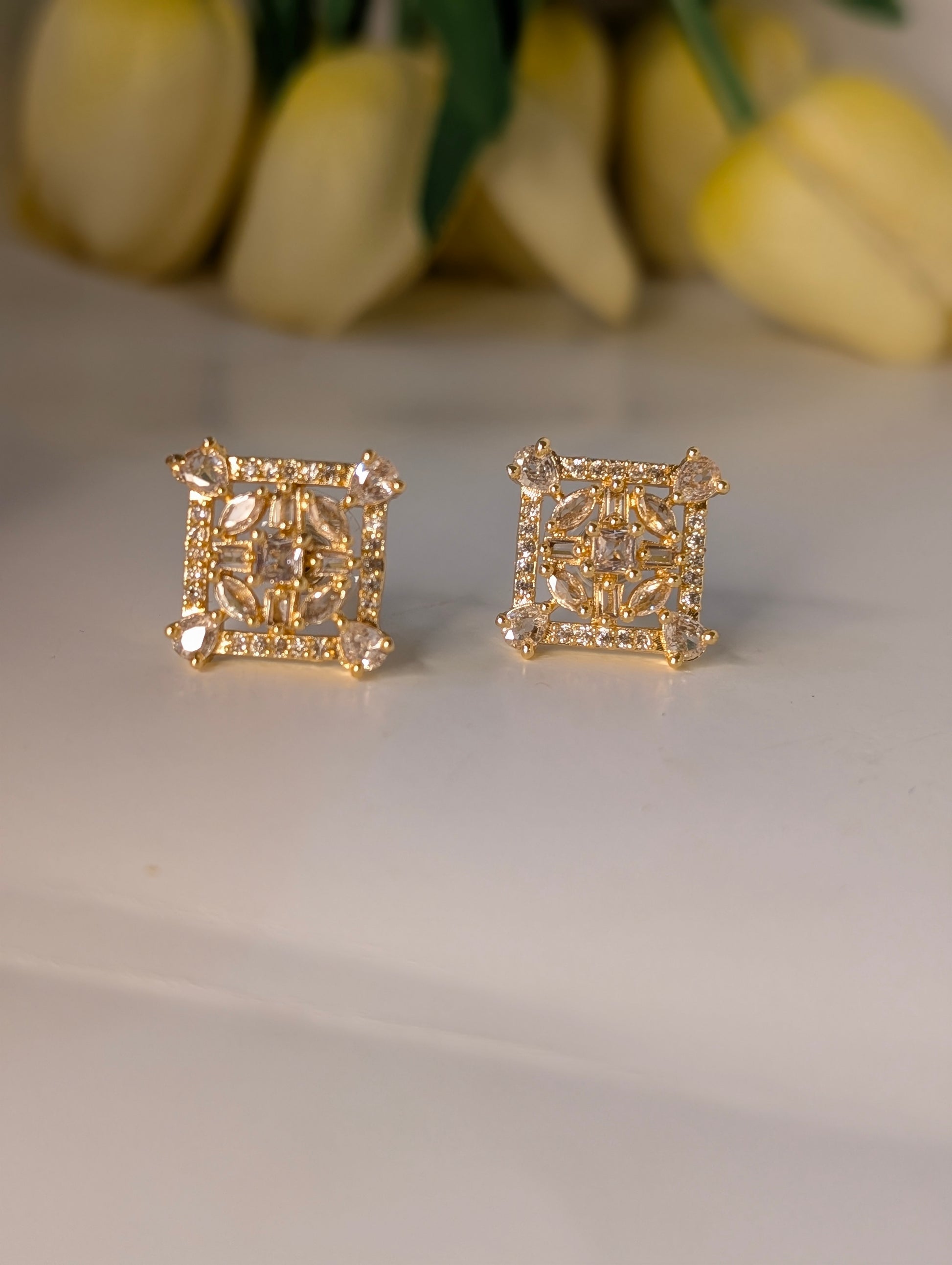 Gold square earrings on a light surface with yellow tulips in the background